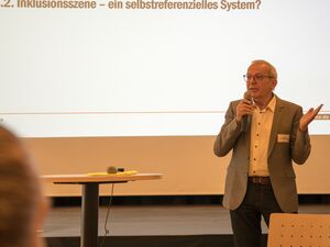 Mann steht vorn und hält Mikro in der Hand. Er hat graue kurze Haare, Brille, er trägt ein graues Jackett, ein weißes Hemd und eine dunkle Hose. Auf der Leinwand hinter ihm steht: 4.2. Inklusionsszene - ein selbstreferenzielles System?