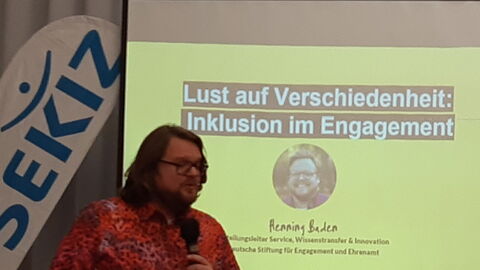 Mann mit einem pink-rot-Leoparden Muster Hemd spricht in ein Mikro hinter ihm läuft eine Präsentation, die er vorstellt. Text: "Lust auf Verschiedenheit: Inklusion im Ehrenamt."