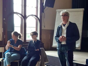 Mann steht vor Flipchart, hat die Hände gefaltet. Er hat graue Haare, Brille, ein weißes Hemd, darüber blaue Jacke und Jeans an. 2 Gebärdendolmetscherinnen sitzen links von ihm.