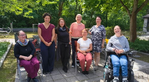 Gruppenfoto: Draußen, Sonne scheint. 7 Frauen. 4 stehend, 2 auf Stühlen sitzend und eine junge Frau im E-Rollstuhl. Alle lächeln.