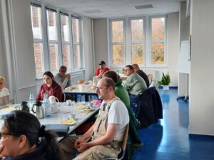 Großer Raum mit vielen Fenstern. Tische mit Getränken, Kuchen und Geschirr. 9 Menschen sitzen verteilt an den Tischen.