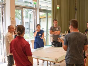 Ein großer Raum, Fensterfront. Um einen Tisch stehen 8 Menschen. Auf dem Tisch liegt ein Flipchart Papier. Frau in blauem Kleids pricht mit Mann gegenüber und gestikuliert mit den Armen.