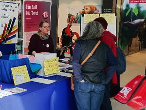 Info-Stand des Netzwerk. Frau mit Brille informiert Interessierte Menschen am Stand.