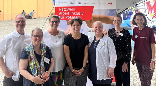 Gruppenfoto: Ein Mann und 6 Frauen. Alle lächeln in die Kamera. Die Gruppe steht vor dem Aufsteller vom Netzwerk Arbeit inklusiv für Potsdam.
