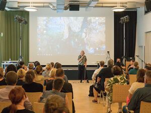 Frau mit Mikrofon und Blindenstock steht vorne. Die hat graue Haare die ihr glatt über die Ohren fall, sie trägt ein grau-schwarzes Pberteil und eine schwarze Hose. Die Teilnehmenden sitzen auf Stühlen. Von weit hinten.