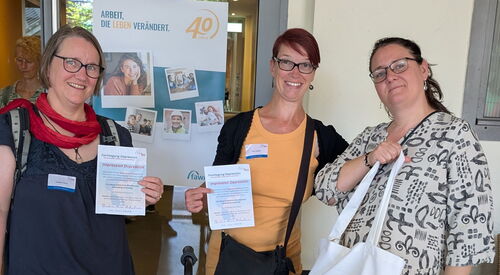 Teamfoto: 3 Frauen vor einem Eingang. alle sommerlich gekleidet. 2 halten das Programm der Tagung in die Kamera.
