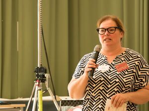 Füllige Frau hält ein Mikro in der rechten Hand und einen Blatt Papier in der linken Hand. Sie hat braune Haare, eine Brille mit schwarzem Rahmen, ein weiß-schwarzes gemustertes kurzärmliges Oberteil. Sie steht vor einen olivgrünen Vorhang und spricht.