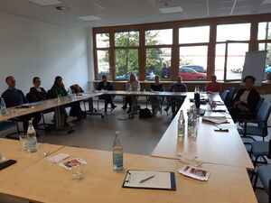 Großer Konferenzraum. Tische, an denen Menschen sitzen.