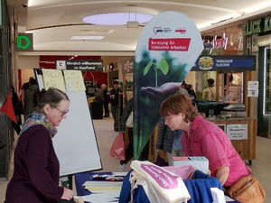 Info- und Mal-Stand des AWO Netzwerks Arbeit inklusiv mit Flipchart, Stiften und Beachflag. Frau mit Brille hinter dem Stand erklärt interessierter Person etwas.
