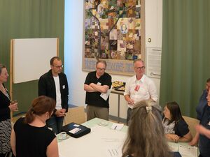 Ca. 10 Personen stehen um einen großen Tisch auf dem ein Flipchart-Papier liegt. Eine Frau sitzt auf einem Stuhl. Eine Gesprächsrunde. Auf dem Papier sind Stichpunkte geschrieben.