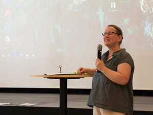 Frau steht an einem Stehtisch. Rechter Arm liegt auf dem Tisch in der linken Hand hält sie ein Mikrofon. Sie lächelt. Sie hat glatte Haare, streng nach hinten gekämmt, eine Brille, sie trägt ein dunkelgrauen T-Shirt mit Polokragen und Knopfleiste.
