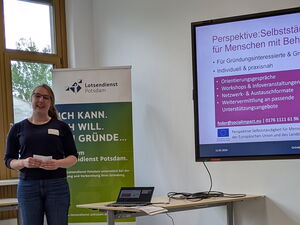 Junge Frau steh links im Bild und spricht. Sie hat einen Zettel in den Händen. An der Wand ein Monitor mit informationen zur Perspektive: Selbstständigkeit für Menschen mit Behinderung.
