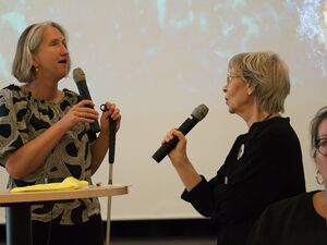 Moderatorin mit Mikro und Blindenstock im Gespräch mit einer Frau die auch ein Mikro in der Hand hält. Frau hat helle kurze Haare und ein schwarzes Kleid an. Die Gebärdendolmetscherin die vorne sitzt guckt konzentriert gerade aus.
