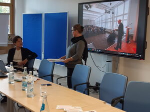 Großer Konferenzraum. Auf einem Bildschirm eine Foto-Präsentation. Davor steht eine Frau und hält eine Rede.