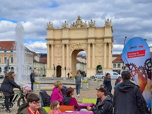 Luisenplatz Potsdam, das Brandenburger Tor im Hintergrund. Vorne Tische und Menschen die Kuchen essen und sich unterhalten. Ein Springbrunnen in der Mitte.