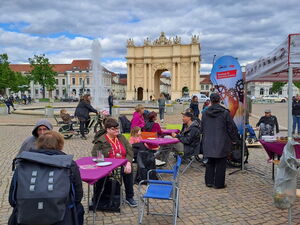 Luisenplatz Potsdam, das Brandenburger Tor im Hintergrund. Vorne Tische und Menschen die Kuchen essen und sich unterhalten. Ein Springbrunnen in der Mitte.