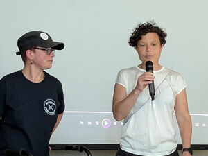 Frau in dunkler Kleidung mit Krüppel gegen Rechts Logo auf dem T-Shirt steht mit Rollator neben einer Frau die in ein Mikrofon spricht. Die Frau trägt ein hellel Oberteil und eine schwarze Hose.