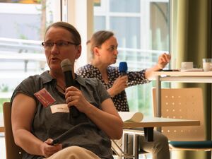 Frau sitzt auf Stuhl, hält ein Mikro in der Hand und spricht. Sie hat glatte Haare, streng nach hinten gekämmt, eine Brille, sie trägt ein dunkelgrauen T-Shirt mit Polokragen und Knopfleiste.
