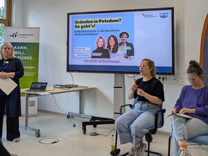 Veranstaltungsraum. Links steht eine Frau und spricht. Rechts im Bild sitzen 2 Gebärden-Dolmetscherinnen. Auf einem Monitor an der Wand dahinter das Veranstaltungs-Bild.