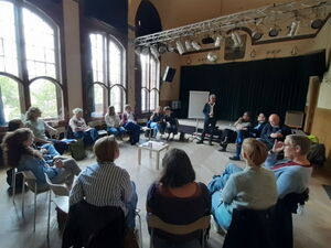 Großer Raum. Großer Stuhlkreis. In der Mitte ein kleiner Tisch. 15 Menschen sitzen auf Stühlen. Mann steht vor Flipchart. Foto von hinten zum Redner.