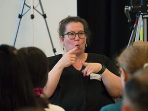 Gebärdendolmetscherin sitzt. Lange dunkle Haare, nach hinten gekämmt, Brille mit schwarzem Rahmen. Sie trägt ein schwarzes T-Shirt. Gespitzter Mund, rechter zeige Finger ausgestreckt vor den Lippen, mit dem linken Zeigefinger hat sie auch ausgestreckt.