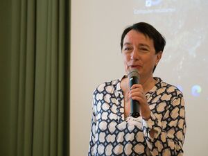 Frau mit Mikrofon in der Hand spricht. Frau trägt kurze schwarze Haare, hat Perlenohrstecker im Ohr. sie ist dezent geschminkt und trägt eine weiße Bluse mit Musterung.