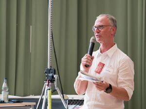 Mann mit Mikro in der rechten Hand und Zetteln in der linken Hand spricht. Er hat kurze graue Haare, trägt eine Brille und ein weißes Hemd. Er steht vor einen olivgrünen Vorhang.