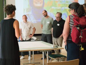 Großer Raum, großer Tisch auf dem ein Flipchart-Papier liegt, Menschen stehen darum herum und diskutieren. Im Hintergrund läuft eine Präsentation einer Ausstellung auf der Leinwand.