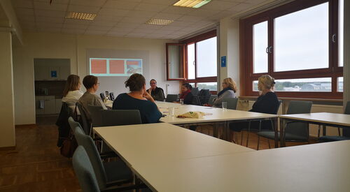 Großer Besprechungsraum. 7 Frauen sitzen um einen großen Tisch. Etwas ist an die Wand projiziert.