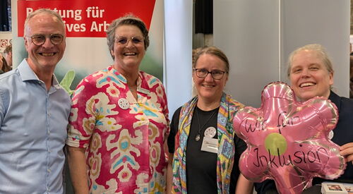 Ein Mann und drei Frauen. Sehr fröhlich an einem Infostand. Frau rechts hält einen rosa Blumenluftballon mit der Aufschrift "Wir für Inklusion."