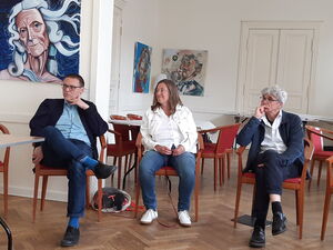 Großer Raum mit Bildern an der Wand. 3 Menschen sitzen auf Stühlen. In der Mitte eine Frau, neben ihr auf dem Boden schläft ein kleiner Hund.