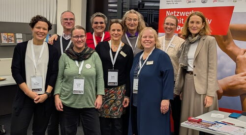 Gruppenbild: 8 Frauen und ein Mann in einem Veranstaltungsraum, mit Roll-Up vom Netzwerk Arbeit inklusiv.