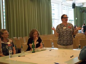 Menschen um einen Tisch, auf dem Tisch, ein Flipchart-Papier mit Stichpunkten. Um den Tisch sitzen Menschen. Eine Frau steht und spricht, sie hat die Hände fragend erhoben.
