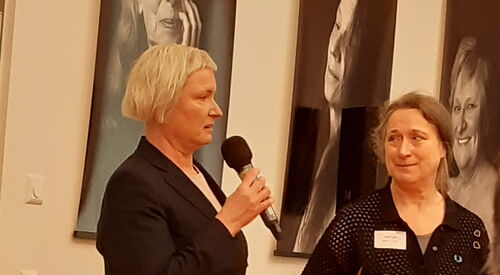 2 Frauen stehen vor großen schwarz-weis- Portraits von Frauen. Sie tragen schwarze Kleidung und eröffnen die Veranstaltung.