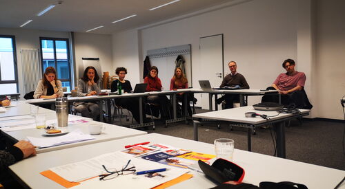 Großer Besprechungsraum. 5 Frauen und zwei Männer sitzen an Tischen. 2 Menschen haben Laptops vor sich die restlichen Zettel und Stifte.