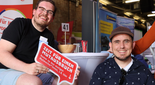 Zwei junge Männer an einem Messestand der AWO. Ein Mann mit dunklen Haaren und Bart, Brille, schwarzes T-Shirt, der andere mit Base Cap. Beide grinsen in die Kamera.