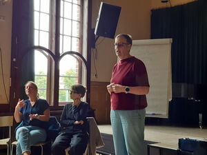 Frau steht vor Flipchart, Sie hat sehr kurze Haare und eine Brille auf, ein dunkelrotes T-Shirt und helle Hosen an. Neben ihr sitzen zwei Gebärdendolmetscherinnen.