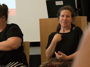 Zwei Frauen (Gebärdendolmetscherinnen) sitzen auf Stühlen. Beide tragen schwarze T-Shirts. Eine der Frauen ist fülliger und dreht sich zur Seite. Die zweite Frau ist schlank und dautet mit dem Zeigefinger der rechten Hand auf sich selbst.