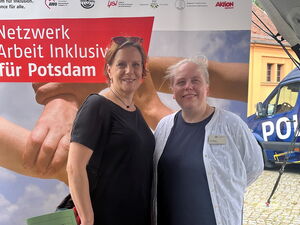 Zwei Frauen vor dem Aufsteller vom Netzwerk Arbeit inklusiv für Potsdam. Die Frauen lächeln in die Kamera.