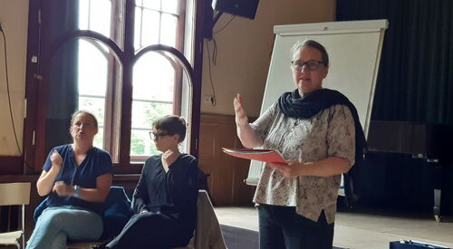 Frau steht vor einem Flipchart und spricht, in der Hand hält sie einen Hefter. Links neben ihr sitzen zwei Frauen in dunkler Kleidung und übersetzen in Gebärdensprache.