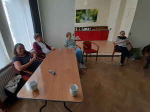 Gesprächsrunde. Großer Raum, Stuhlkreis, ein Tisch links, mit zwei Tassen und 2 Gläsern. Vier Personen sitzen im großen Raum.