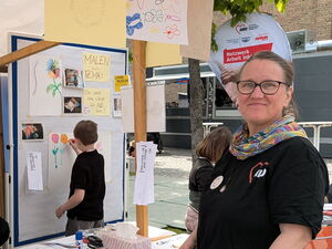 Eine Frau mit Brille steht lächeln an einem Kreativ Stand zum Malen.  Junge malt im Hintergrund Blumen.
