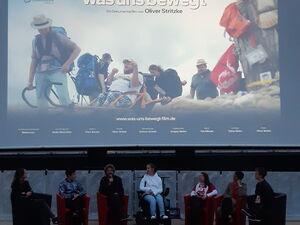 Diskussionsrunde: 6 Menschen sitzen in Sesseln, in der Mitte eine Rollstuhlfahrerin im E-Rollstuhl. Das Mikrofon hat ein Mann im dritten Sessel. Darüber die Kinoleinwand mit dem Film-Titelbild.