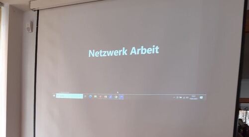 Tisch mit Flyern, Darüber Leinwand auf die Zoom Meeting projiziert wird. Netzwerk Arbeit steht dort.