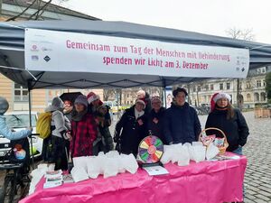 Pavillon, Tisch vorne auf dem kleine Papiertüten, kleines Glücksrad, ein Körbchen mit Süßigkeiten steht. Menschen stehen im Pavillon. Einige lächeln in die Kamera.