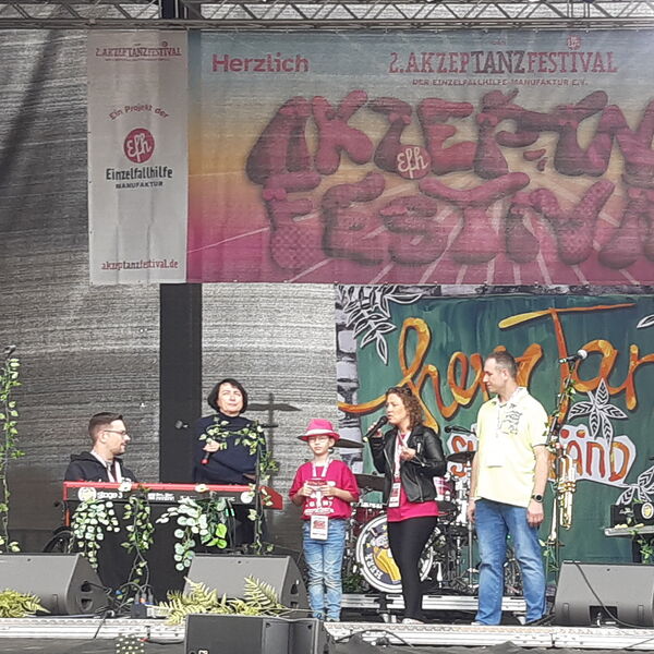 Auf der Bühne stehen 5 Personen, Rollstuhlfahrer, zwei Frauen, ein Junge mit pinker Festival Kleidung und Hut, Moderatorin mit Mikro und ein Mann im gelben T-Shirt.