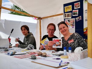 3 Personen im AWO Stand. Junger Mann an Button-Maschine und zwei Frauen die lachend Info-Flyer und Broschüren in die Kamera halten.