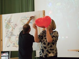 Frau bläst einen roten herzförmigen Luftballon auf. Sie hat graue Haare die ihr glatt über die Ohren fallen, sie trägt ein grau-schwarzes Oberteil. Mann im schwarzen T-Shirt zeichnet noch das Abschluss Bild an das Flipchart-Papier.
