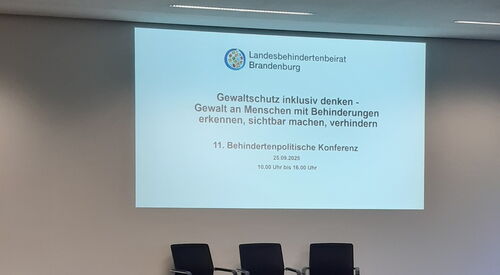 Leerer Konferenzraum, drei Stühle auf einem Podium. Darüber mit Beamer an die Wand projiziert. "Gewaltschutz inklusiv denken - Gewalt an Menschen mit Behinderung erkennen, sichtbar machen, verhindern.