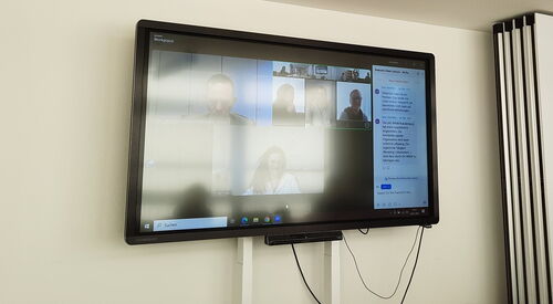 Monitor an der Wand. Ein Online Meeting. Mehrere Menschen sind zugeschaltet.